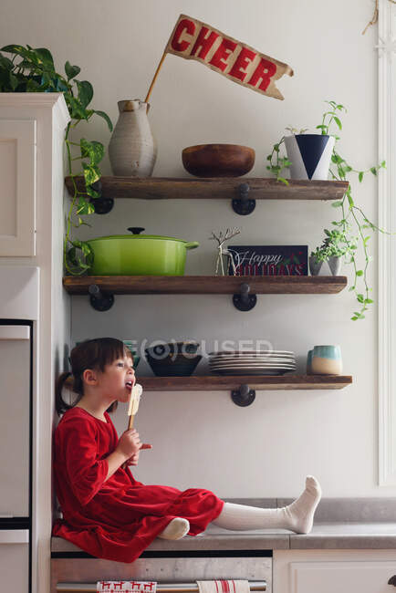 Girl sitting on kitchen counter with a spatula eating frosting — Stock Photo