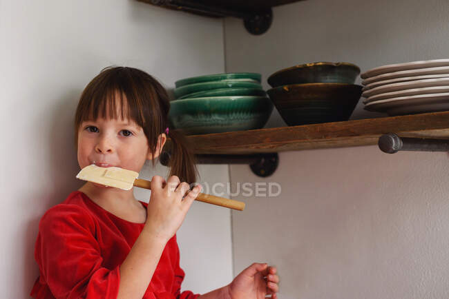 Menina sentada no balcão da cozinha com uma espátula comendo cobertura — Fotografia de Stock