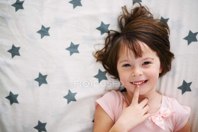 Blick auf lächelndes Mädchen, das mit dem Finger auf den Lippen im Bett liegt — Stockfoto