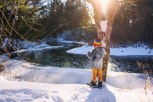 Ragazzo con le ciaspole in piedi vicino al fiume in inverno — Foto stock