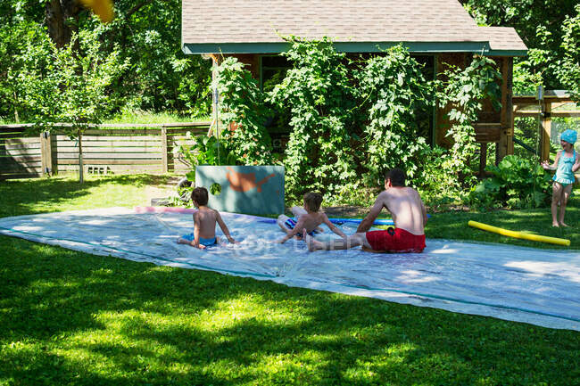 Padre y tres niños jugando en un resbalón y deslizarse en el jardín - foto de stock