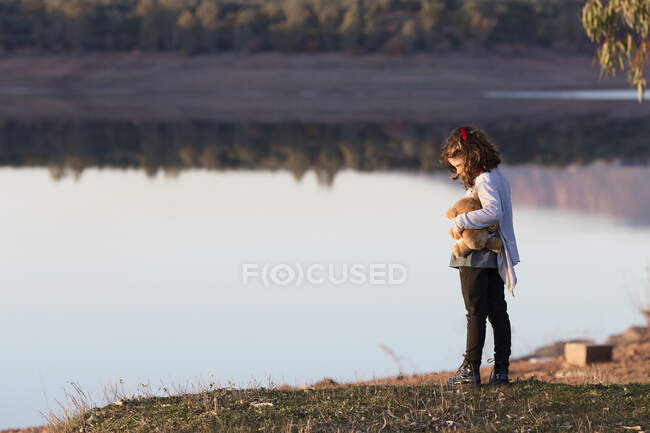 Chica de pie junto a un lago con su oso de peluche - foto de stock