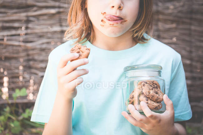 Menino comer biscoito de chocolate enquanto segurava um frasco de biscoitos — Fotografia de Stock