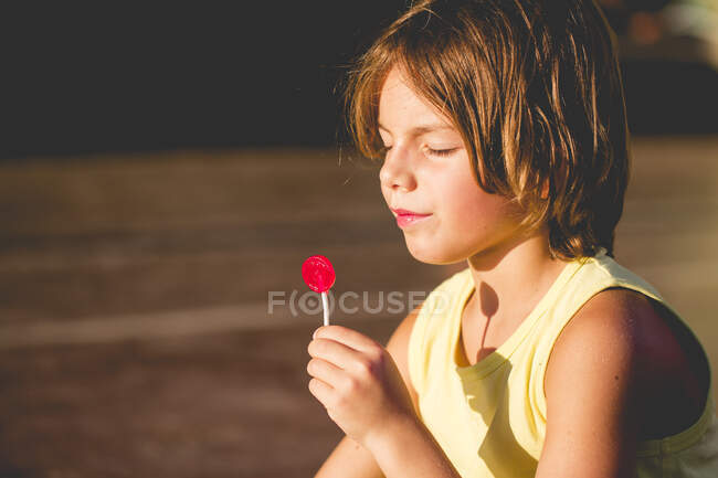 Niño sentado en el sol comiendo una piruleta - foto de stock
