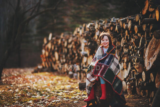 Girl sitting by log pile wrapped in patchwork quilt — Stock Photo