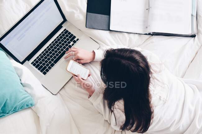 Дівчина-підліток лежить в ліжку, використовуючи свій ноутбук і мобільний телефон — стокове фото