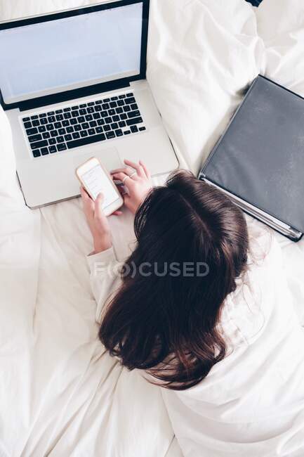 Adolescente acostada en la cama usando su computadora portátil y teléfono móvil - foto de stock