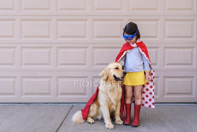 Girl dressed as a superhero standing by the garage with her golden retriever dog — Stock Photo