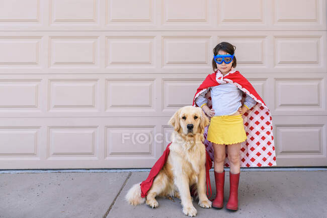 Fille habillée en super-héros debout près du garage avec son chien golden retriever — Photo de stock
