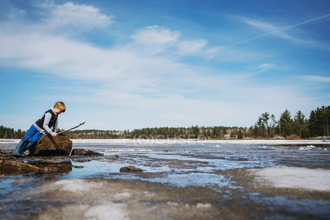 Boy standing by a frozen lake holding a stick — Stock Photo
