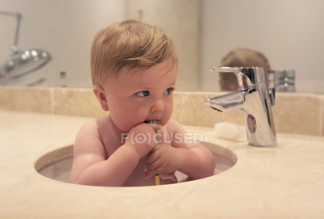 Baby boy sitting in a sink brushing his teeth — Stock Photo