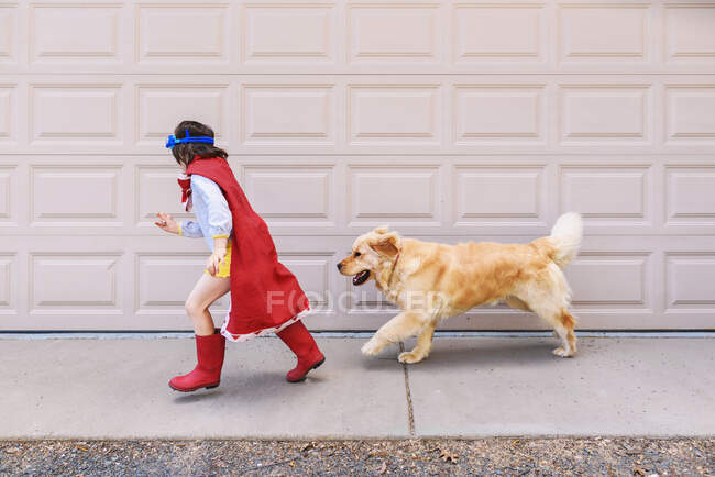 Girl running past a garage door dressed as a superhero with her dog — Stock Photo