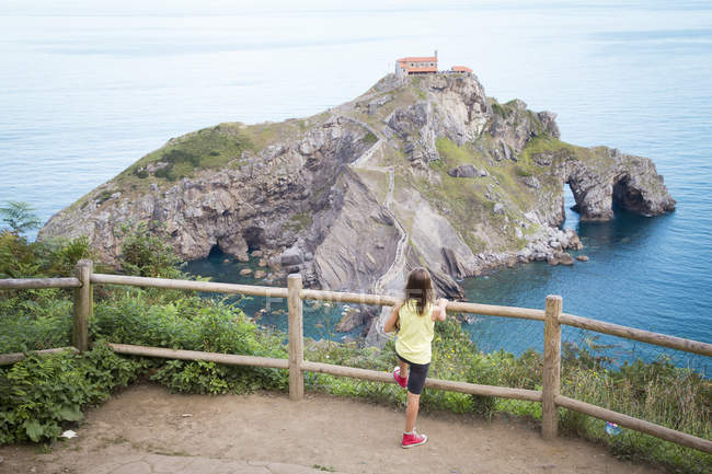 Girl standing by a fence looking at a hermitage on San Juan de Gaztelugatxe, Bermeo, Vasco, Spain — Stock Photo