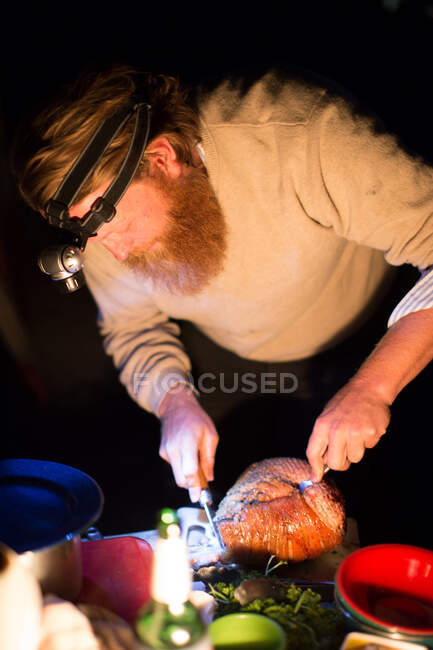 Man carving a roasted ham at night at a campsite — Stock Photo