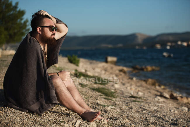 Man sitting on beach wrapped in a towel with his hand in his hair — Stock Photo