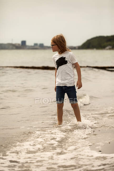 Menino de pé na praia no surf olhando para o mar — Fotografia de Stock