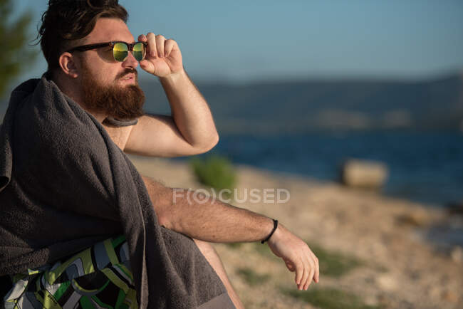 Portrait d'un homme assis sur la plage avec sa main sur ses lunettes de soleil — Photo de stock