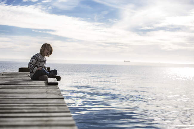 Niño sentado en un muelle de madera junto al mar, Aarhus, Dinamarca - foto de stock