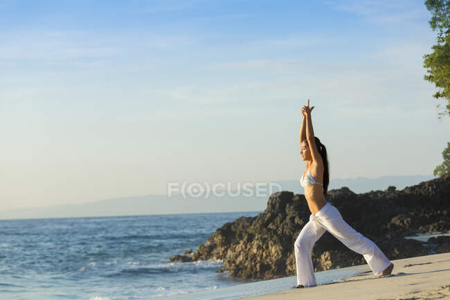 A young asian woman is doing yoga at a white sandy beach in Bali. She is wearing long white pants and a bikini top. — Stock Photo