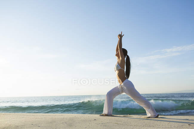 Una joven asiática está haciendo yoga en una playa de arena blanca en Bali. Lleva pantalones blancos largos y un top de bikini.. - foto de stock