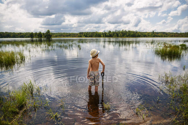 Young boy wading into peaceful lake with reflection of sky and clouds with a shovel — Stock Photo