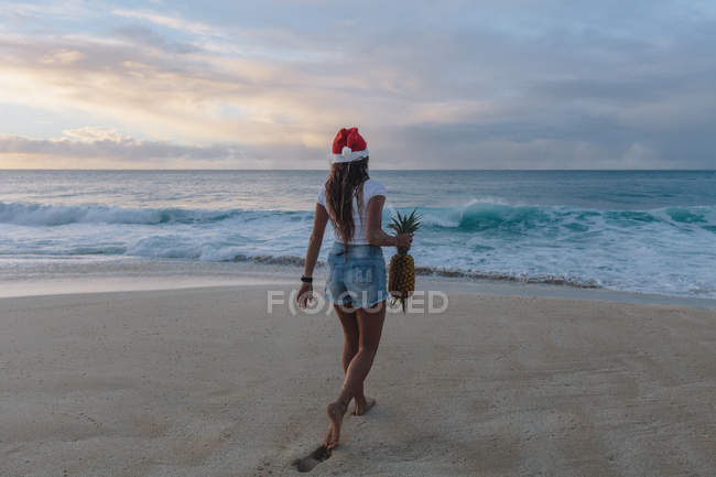 Woman wearing a Christmas Santa hat walking on beach carrying a pineapple, Haleiwa, Hawaii, America, USA — Stock Photo