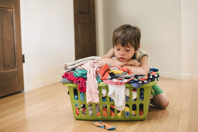 Cansado Niño sentado en el suelo apoyado en una lavandería disfrutado lleno de ropa - foto de stock
