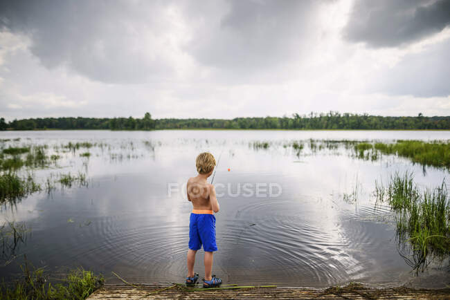 Молодий хлопчик ловить рибу в спокійному озері з відображенням неба і хмар. — стокове фото