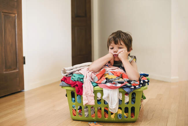 Garoto cansado sentado no chão apoiado em uma lavanderia repleta de roupas — Fotografia de Stock