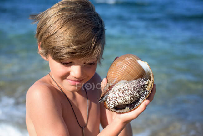 Ragazzo sorridente sulla spiaggia che tiene una conchiglia di fronte al mare — Foto stock