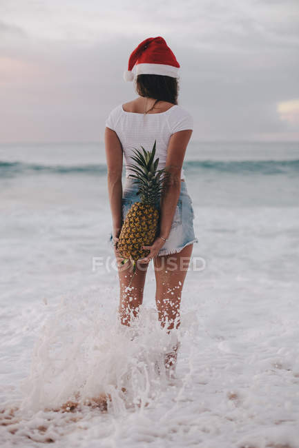 Donna che indossa un cappello di Babbo Natale in piedi in oceano surf tenendo un ananas dietro la schiena, Haleiwa, Hawaii, America, Stati Uniti d'America — Foto stock