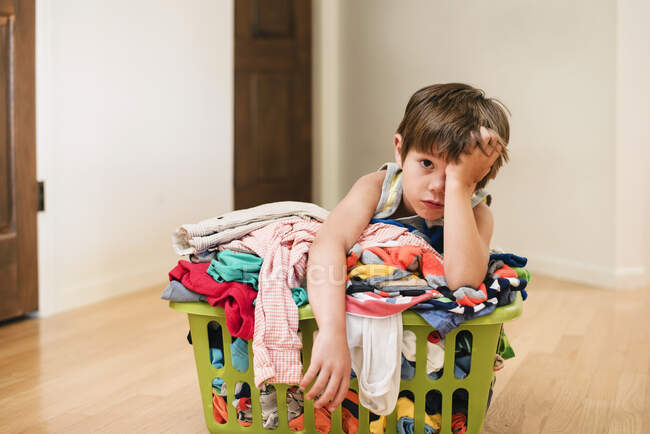 Menino sentado no chão encostado em uma lavanderia repleta de roupas — Fotografia de Stock