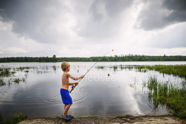 Jeune garçon tenant des poissons près d'un lac paisible avec reflet du ciel et des nuages — Photo de stock