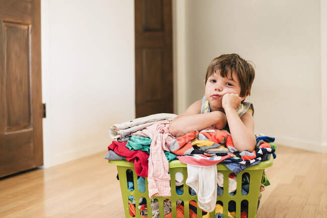 Menino sentado no chão encostado em uma lavanderia repleta de roupas — Fotografia de Stock