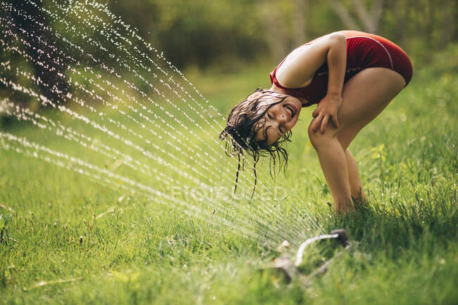 Chica joven jugando en un aspersor en el patio trasero - foto de stock