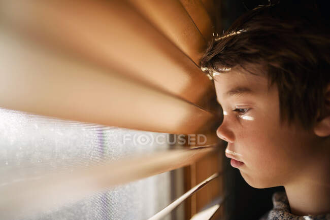 Garçon debout près d'une fenêtre regardant à travers les stores — Photo de stock