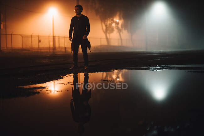 Hombre parado en la carretera junto a un charco de agua por la noche, California, América, EE.UU. - foto de stock