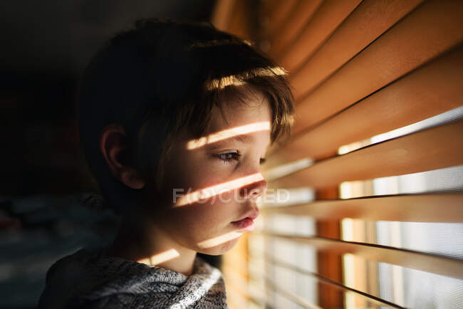 Garçon debout près d'une fenêtre regardant à travers les stores — Photo de stock