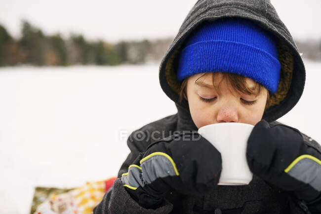 Niño de pie en la nieve bebiendo chocolate caliente - foto de stock
