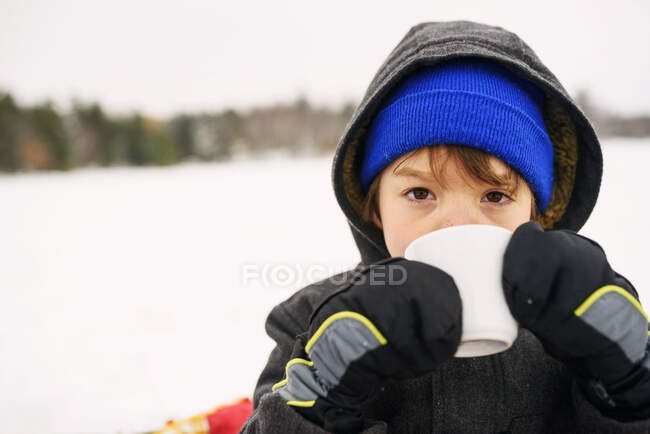 Хлопчик стоїть на снігу п'є гарячий шоколад — стокове фото