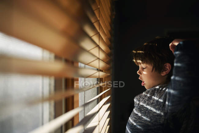 Niño de pie junto a una ventana bostezando y estirando - foto de stock