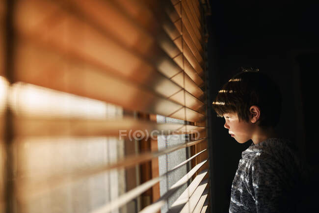 Rapaz parado junto a uma janela a olhar através de persianas — Fotografia de Stock
