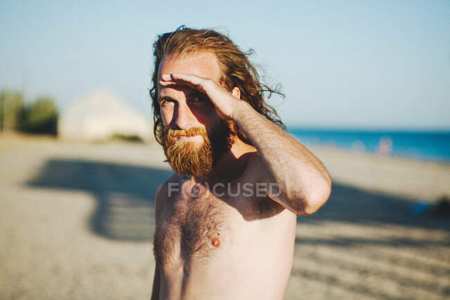 Мужчина с длинными волосами и бородой стоит на пляже, защищая глаза от солнца. — стоковое фото