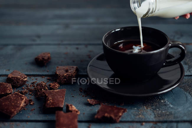 Mão derramando leite em uma xícara de café com chocolate escuro — Fotografia de Stock