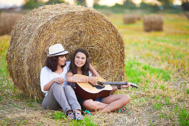 Madre e hija sentadas junto a una paca de heno tocando la guitarra, Bulgaria - foto de stock
