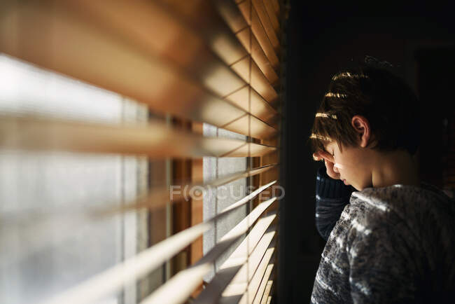 Niño cansado de pie junto a una ventana frotándose los ojos - foto de stock