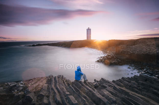 Femme assise sur des rochers au bord de la mer regardant le coucher du soleil, région du Nord-Ouest, Islande — Photo de stock