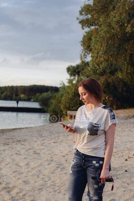 Жінка стоїть на пляжі дивиться на свій мобільний телефон — стокове фото