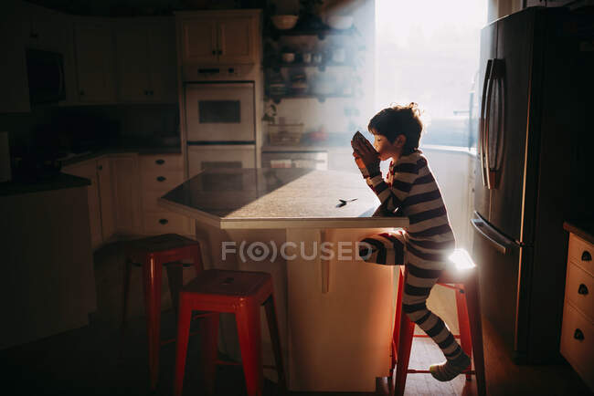 Boy sitting in kitchen eating his breakfast in morning light — Stock Photo