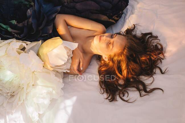 Overhead view of a woman lying on a bed amongst giant artificial peony flowers — Foto stock